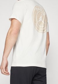 T-shirt en coton blanc présentant un grand logo circulaire beige au dos avec l'inscription "PARIS SAINT-GERMAIN" et une silhouette Jumpman. Manches courtes.