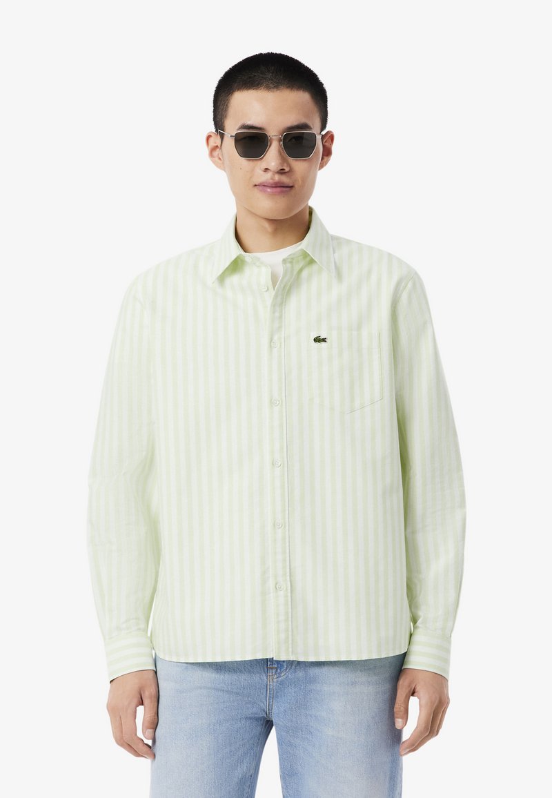 Uomo che indossa una camicia a righe verticali bianche e verde chiaro, jeans azzurri e occhiali da sole scuri, in piedi davanti a uno sfondo bianco.