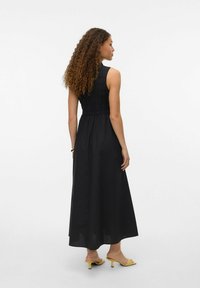 Robe maxi noire avec un buste ajusté mettant en valeur une texture froncée, sans manches, et un ourlet fluide. Associée à des talons jaunes.