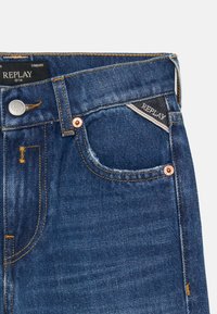 Trapke od trapera u tamnoplavoj boji s izblijedjelom teksturom. Imaju srebrni gumb, bakrene zakovice i oznaku s logotipom na prednjem džepu.