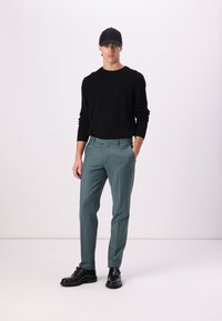 Suéter negro de punto, pantalones ajustados color verde azulado y zapatos de cuero negros. El modelo está de pie con una gorra, en un fondo claro y sencillo.