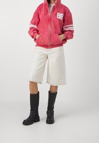 Sudadera roja con cremallera y logo 86 blanco con rayas, combinada con pantalones cortos de mezclilla beige y botas negras altas hasta la rodilla con hebillas.