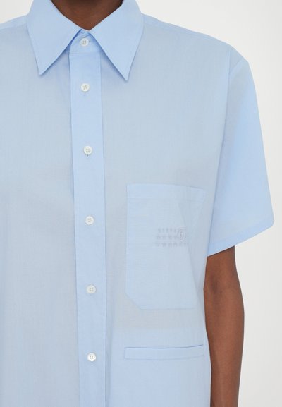 Camicia azzurro chiaro a maniche corte realizzata in tessuto morbido, con una chiusura frontale a bottoni, una tasca sul petto e numeri in rilievo sulla tasca.