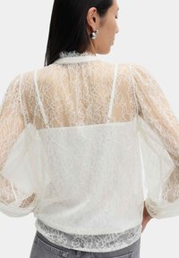 Blusa bianca in pizzo con un design trasparente, delicati motivi floreali e polsini elasticizzati, abbinata a un sottogonna uniforme.