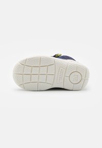 Navy blue sneaker with a thick white rubber sole, textured tread, and green accent. Velcro strap closure. Durable textile upper.