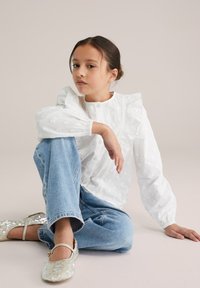 Blouse blanche texturée avec manches volantées, associée à un jean en denim bleu clair. Comprend également des ballerines à sequins. Design simple et épuré.