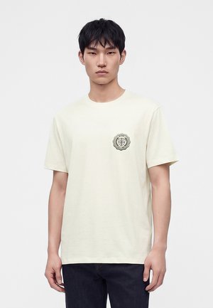 Jeune homme portant un t-shirt beige clair uni à manches courtes avec un petit emblème rond sur la poitrine gauche, debout devant un fond uni.