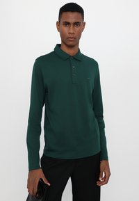 Langermet polo-skjorte i dyp grønn. Har krage, tre knapper i plakket og logodetalj. Materialet virker glatt og teksturert.