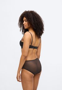 Μαύρο σουτιέν από δαντέλα με ρυθμιζόμενες τιράντες, συνδυασμένο με ημιδιάφανα μαύρα ψηλόμεσα εσώρουχα με κυματιστά τελειώματα και μαλακή υφή.
