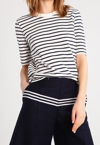 Žena v bílo-námořnickém pruhovaném krátkém rukávu, zastrčeném do vysokých námořnických kalhot s odpovídajícími horizontálními pruhy.