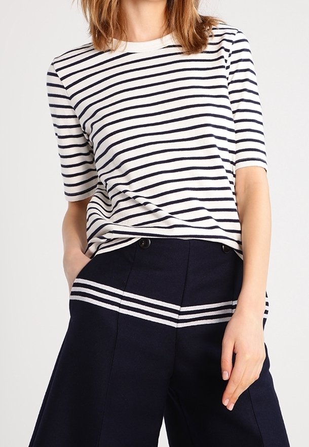 Žena v bílo-námořnickém pruhovaném krátkém rukávu, zastrčeném do vysokých námořnických kalhot s odpovídajícími horizontálními pruhy.