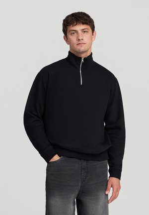 Maglione nero a collo alto con zip a un quarto, tessuto morbido, vestibilità comoda e polsini a coste, abbinato a jeans grigi. Design semplice e neutro.