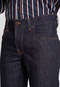 Mörkblå denimjeans med orange sömmar, rak passform, femficksdesign och metallknapp vid midjan.