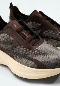 Een paar grijze en donkerbruine gebreide sneakers met gevoerde crèmewitte zolen en oranje "OYSH" branding op de tong en zijkant.