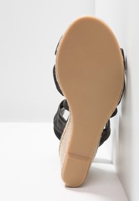 Sandale compensée avec semelle en caoutchouc marron clair, brides noires à la cheville et talon espadrille tressé, debout sur fond blanc.