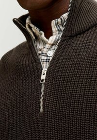 Brauner Strickpullover mit einem Reißverschlusskragen, der ein strukturiertes Muster und einen Reißverschluss aus rostfreiem Stahl aufweist, über einem karierten Hemd getragen.