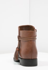 Bottine en cuir marron avec une sangle tressée et un détail de gland. Semelle noire plate, bout arrondi et texture lisse. Vue de dos.