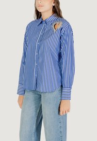 Camicia button-up a righe blu e bianche con un'apertura a forma di cuore e un'imbellitura decorativa sulla spalla sinistra. Tessuto di cotone, maniche lunghe.