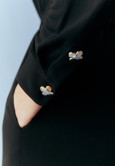 Black blouse sleeve with textured fabric, featuring gold and silver floral-shaped buttons at the cuff. Clean lines and fitted design.