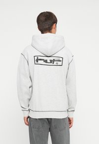 Grå hettegenser med en stor svart "huf" logo på baksiden, samt ribbestrikkede mansjetter og nederkant. Mykt materiale med synlige sømmer.