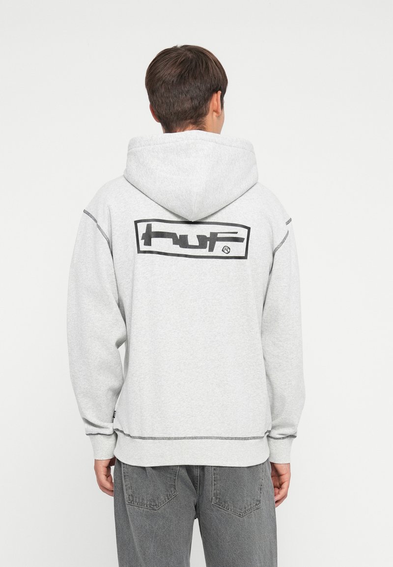Grå hettegenser med en stor svart "huf" logo på baksiden, samt ribbestrikkede mansjetter og nederkant. Mykt materiale med synlige sømmer.