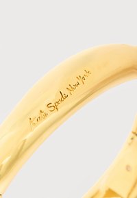 Βραχιόλι σε χρυσό τόνο με εγχάρακτο λογότυπο "kate spade New York" σε λείο, κυρτό επίπεδο πάνω σε απλό ανοιχτό φόντο.