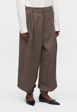 Pantalon marron à jambes larges avec poignets élastiques porté avec une chemise blanche à fines rayures et des ballerines noires pointues.