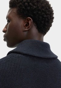 Donker, gestructureerd wollen jack met een brede kraag, met een close-up van de achterkant. Geweven patroon zichtbaar. Korte, krullende haren op de achtergrond.