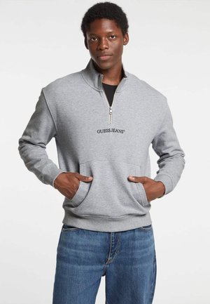 Grauer Reißverschluss-Sweatshirt mit hohem Kragen, ausgestattet mit einer Fronttasche und besticktem "GUESS JEANS"-Logo. Kombiniert mit blauer Denim-Jeans.