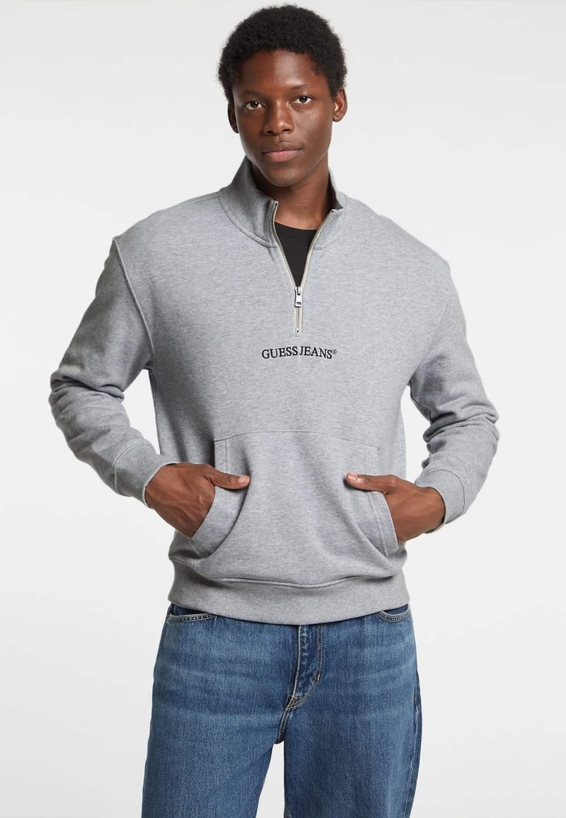 Pelēks rāvējslēdzēja džemperis ar augstu apkakli, ar priekšējo kabatu un izšūtu "GUESS JEANS" logo. Saskaņots ar ziliem džinsiem.