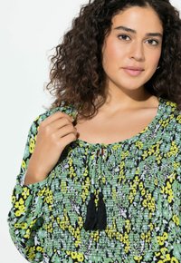 Blusa floreale verde con accenti gialli e neri. Presenta un collo arricciato con frange nere. Realizzata in un tessuto leggero e testurizzato.