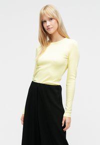 Sárga hosszú ujjú felső bordázott anyagból, fekete pliszírozott szoknyával párosítva. A ruházat kerek nyakkivágással és testhez simuló kialakítással rendelkezik.