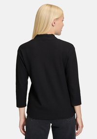 Donna bionda di spalle, che indossa un top nero a maniche lunghe con colletto alto e pantaloni neri, su uno sfondo chiaro e semplice.