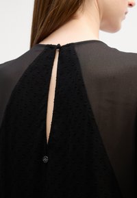 Rochie neagră cu un spate texturat, decorat cu modele circulare, o deschidere în formă de cheie și o mică închidere cu nasture la guler.