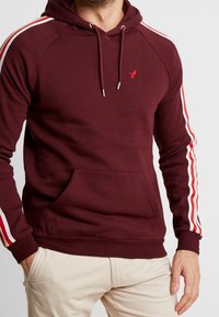 Hombre vestido con sudadera color burdeos con rayas blancas y rojas en las mangas y pantalones beige, con las manos descansando cerca de los bolsillos.