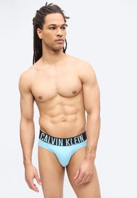 Άνδρας χωρίς πουκάμισο με dreadlocks, φορώντας ανοιχτό μπλε εσώρουχο Calvin Klein, στέκεται μπροστά σε απλό φωτεινό φόντο, κοιτώντας προς τα αριστερά του.