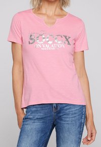 Ljusrosa t-shirt med V-hals, med silverfärgad grafik som står "SOCcx ON VACATION" och en avslappnad passform. Mjuk bomullsmaterial.