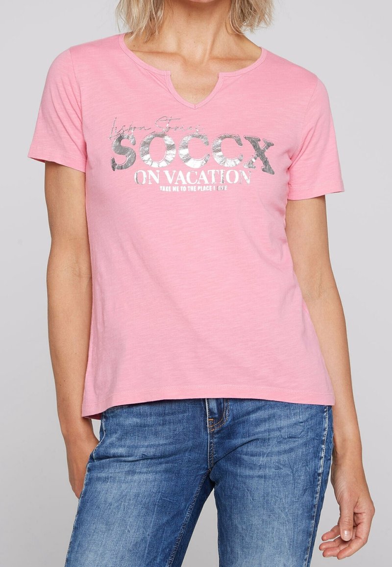 Ljusrosa t-shirt med V-hals, med silverfärgad grafik som står "SOCcx ON VACATION" och en avslappnad passform. Mjuk bomullsmaterial.