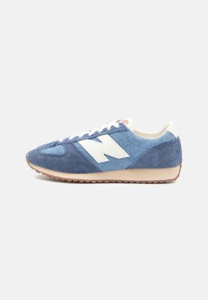 Blauer und marineblauer Veloursleder-Sneaker mit weißem "N"-Logo, beiger Sohle und weißen Schnürsenkeln auf hellem einfarbigem Hintergrund.