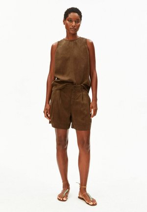 Frau steht, trägt ein braunes ärmelloses Oberteil und passende Shorts mit braunen und grünen Sandalen, vor einem schlichten weißen Hintergrund.