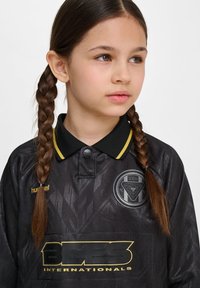 Svart tröja med geometriska mönster, gula accenter och krage i svart. Har en logotyp och varumärkesnamn framtill. Slät textur.