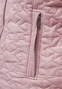 Růžová prošívaná tkanina bundy s diamantovým vzorem. Má na boku kapsu na zip s kovovým kováním. Hladký, texturovan�ý povrch.