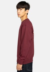 Bordeaux sweatshirt met lange mouwen en geribbelde manchetten, voorzien van een wit grafisch ontwerp op de rug, gedragen over donkere jeans.