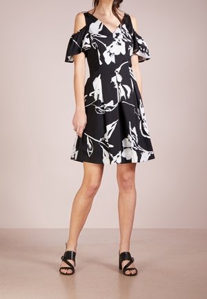 Woman wearing black cold-shoulder dress with large white floral print and black open-toe block heel sandals, standing on beige floor.