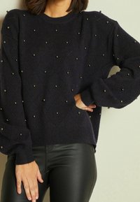 Granatowy sweter z dzianiny z teksturowanym wzorem w diamenty, ozdobiony małymi okrągłymi elementami. Sweter ma prążkowane mankiety i dół, zestawiony z czarnymi obcisłymi spodniami.
