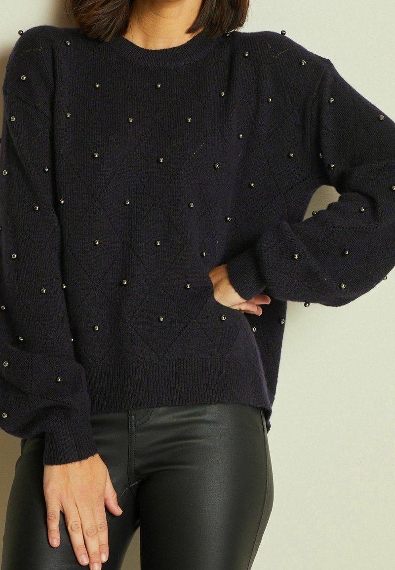 Granatowy sweter z dzianiny z teksturowanym wzorem w diamenty, ozdobiony małymi okrągłymi elementami. Sweter ma prążkowane mankiety i dół, zestawiony z czarnymi obcisłymi spodniami.