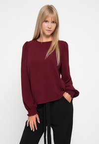 Top burdeos com um acabamento texturizado, decote redondo e mangas bufantes. Apresenta punhos franzidos e é combinado com calças pretas de cintura alta.