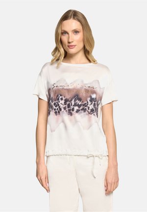 Femme aux cheveux blonds portant un T-shirt blanc ample avec un imprimé léopard et le texte « Parfois, moins c'est plus », associé à un pantalon blanc.