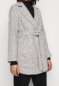 Abrigo de punto gris con diseño envolvente, solapas anchas y cierre con lazo. Tejido texturizado y bolsillos laterales; se lleva sobre una blusa negra.
