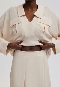 Ljusbeige blus med V-ringning, oversized krage och sidofickor, i kombination med matchande högmidjade byxor med en slät textur.